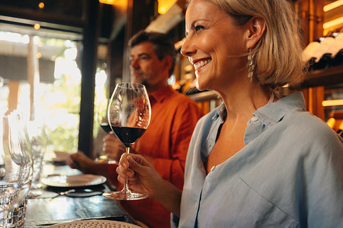 Smiling woman enjoying a glass of wine at a luxury bar with mature man in the background