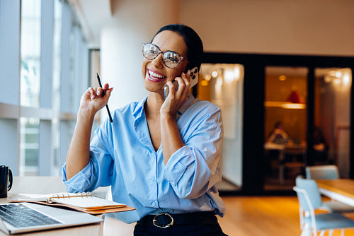 Smiling businesswoman talking on phone at desk with laptop and notes