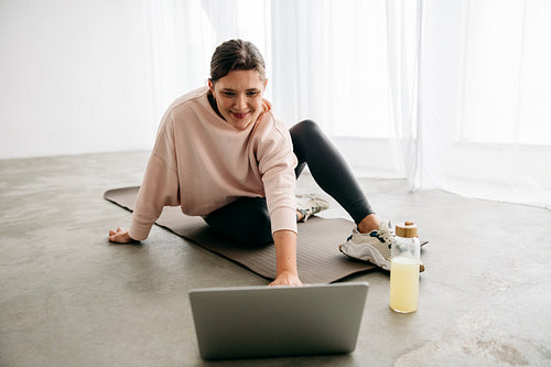 Woman using laptop for online fitness session on yoga mat at home