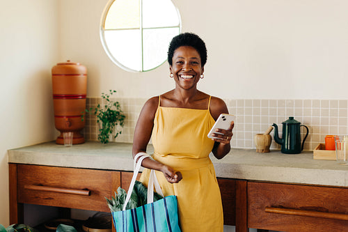 Brazilian woman smiling and holding a vegetable bag with mobile phone in kitchen at home