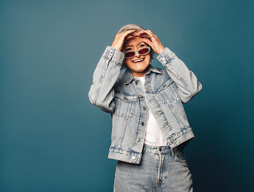 Cheerful senior woman having fun in blue denim outfit and glasses on grey background