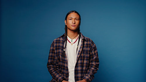 Young Indigenous man with braids and necklace against a blue backdrop