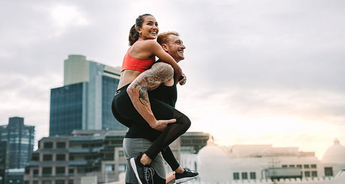 Cheerful man carrying a fitness woman on his back walking on rooftop
