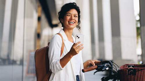 Smiling woman with smartphone and backpack standing by bike with basket on urban street