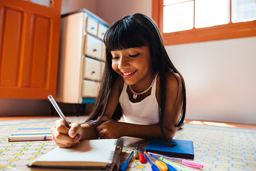 Young girl writing in a notebook on the floor with colorful pens