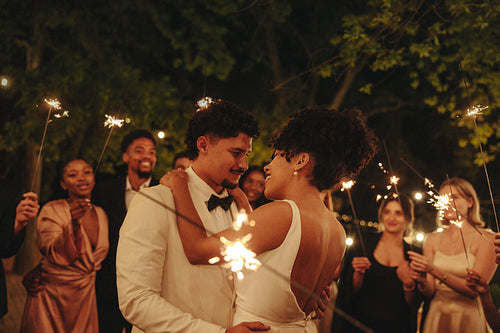 Joyful newlywed couple dancing surrounded by guests holding sparklers at night