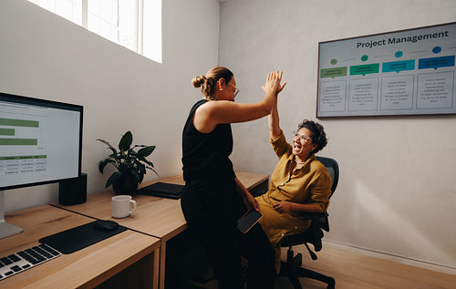 Two women celebrate teamwork with a high-five in a modern office setting