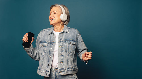 Senior woman dances to music with smartphone and headset against blue background