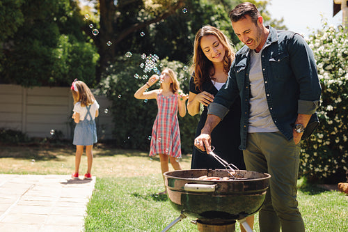 Couple cooking grilled food in backyard with children playing