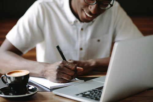 Man writing notes sitting at a coffee shop with a laptop on the table