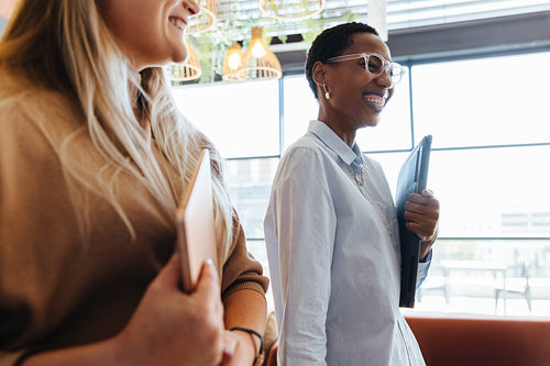 Two women smiling indoors while carrying notebooks and folders