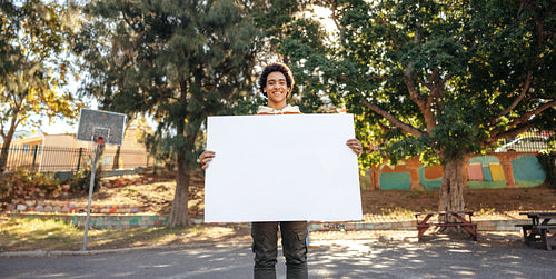 Cheerful teenage boy holding a blank placard in an urban park