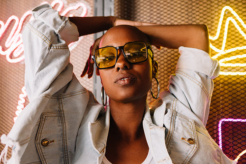 Confident young woman with stylish sunglasses standing in front of neon lights, urban fashion