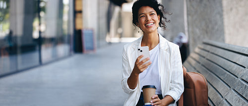 Happy woman sitting on bench texting on smartphone and drinking coffee outdoors