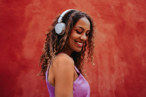 Relaxed woman listens to music on headphones outdoors by a red wall