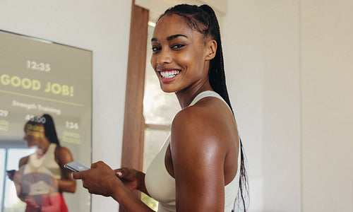 Happy young woman using a smartphone and a smart mirror for fitness
