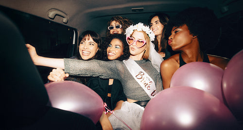 Young women in the back of a taxi taking a selfie and celebrating a bachelorette party with balloons