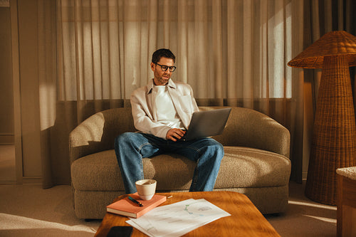 Man works on laptop in office workspace from a sofa with notebook and coffee