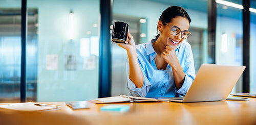 Smiling woman enjoying coffee while working on a laptop at her desk
