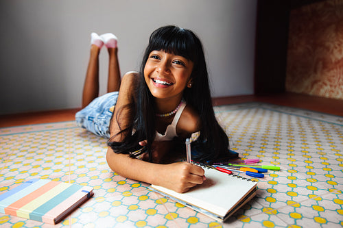 Smiling young girl drawing on a notebook while lying on colorful floor