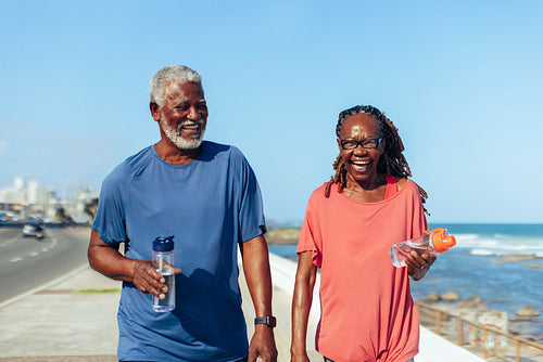 Joyful older couple taking a walk on the seaside promenade holding water bottles