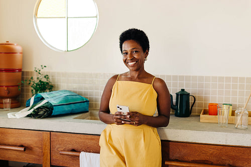 Cheerful brazilian woman smiling and using smartphone in kitchen