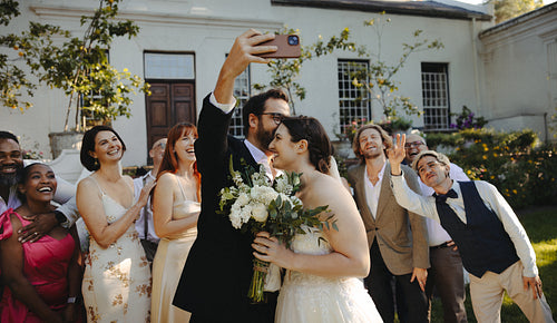 Bride and groom taking a joyful group selfie with wedding guests outdoors