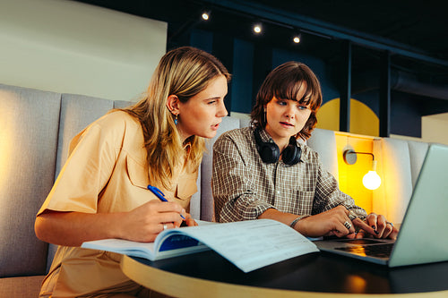 Female students collaborating on a project, studying together with a laptop and textbooks in a modern study space