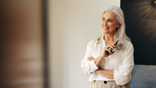 Thoughtful senior woman holding mobile phone indoors