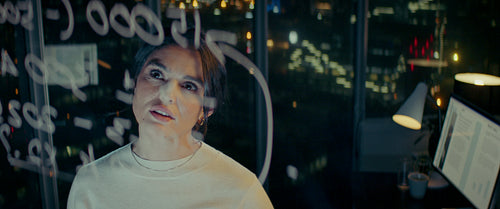 Young woman working late at night, solving equations on a glass partition in her office