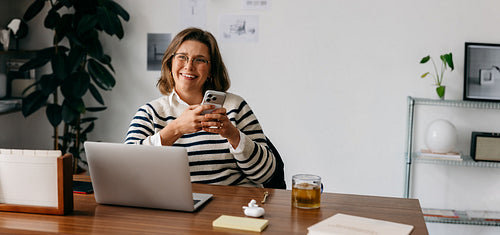 The woman checks her smartphone beside a laptop in a busy home office