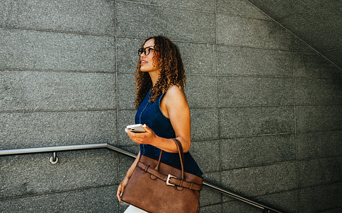Curly-haired business woman walks up stairs with smartphone during urban commute