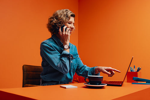 Business owner talking on smartphone while pointing at laptop in a bright orange office