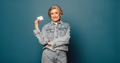 Senior woman with happy smile and credit card standing against blue denim background