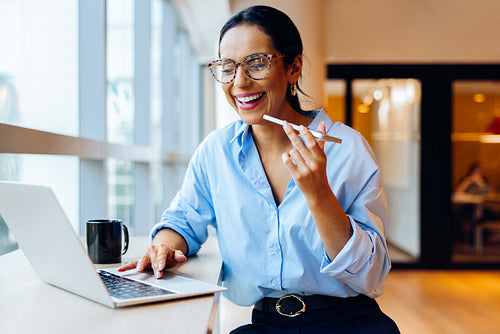 Smiling woman using laptop and speaking into a smartphone at a desk