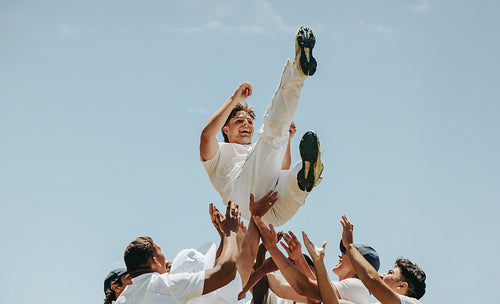 Cricketers lifting a teammate in celebration during a game under a clear sky