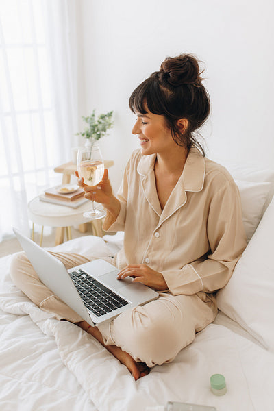 Woman sitting on bed with laptop and enjoying wine