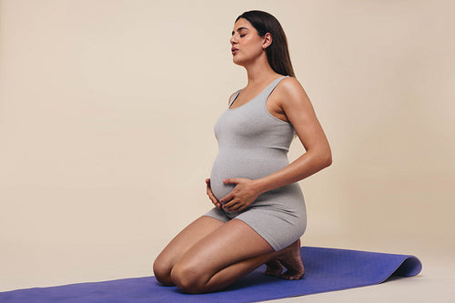 Happy pregnant woman kneeling on the floor in a studio, smiling at her  beautiful baby bump. Young mom-to-be wearing lingerie, cherishing the life  growing inside her with maternal love. stock photo